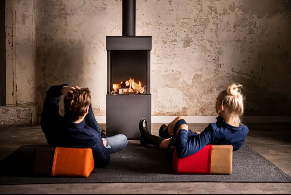 Ungt par som slapper av på fargerike gulvputer foran en moderne høy svart frittstående gasspeis med varme realistiske flammer, i et stilfullt industrielt loft med rå betongvegger – koselig moderne livsstil stock-foto