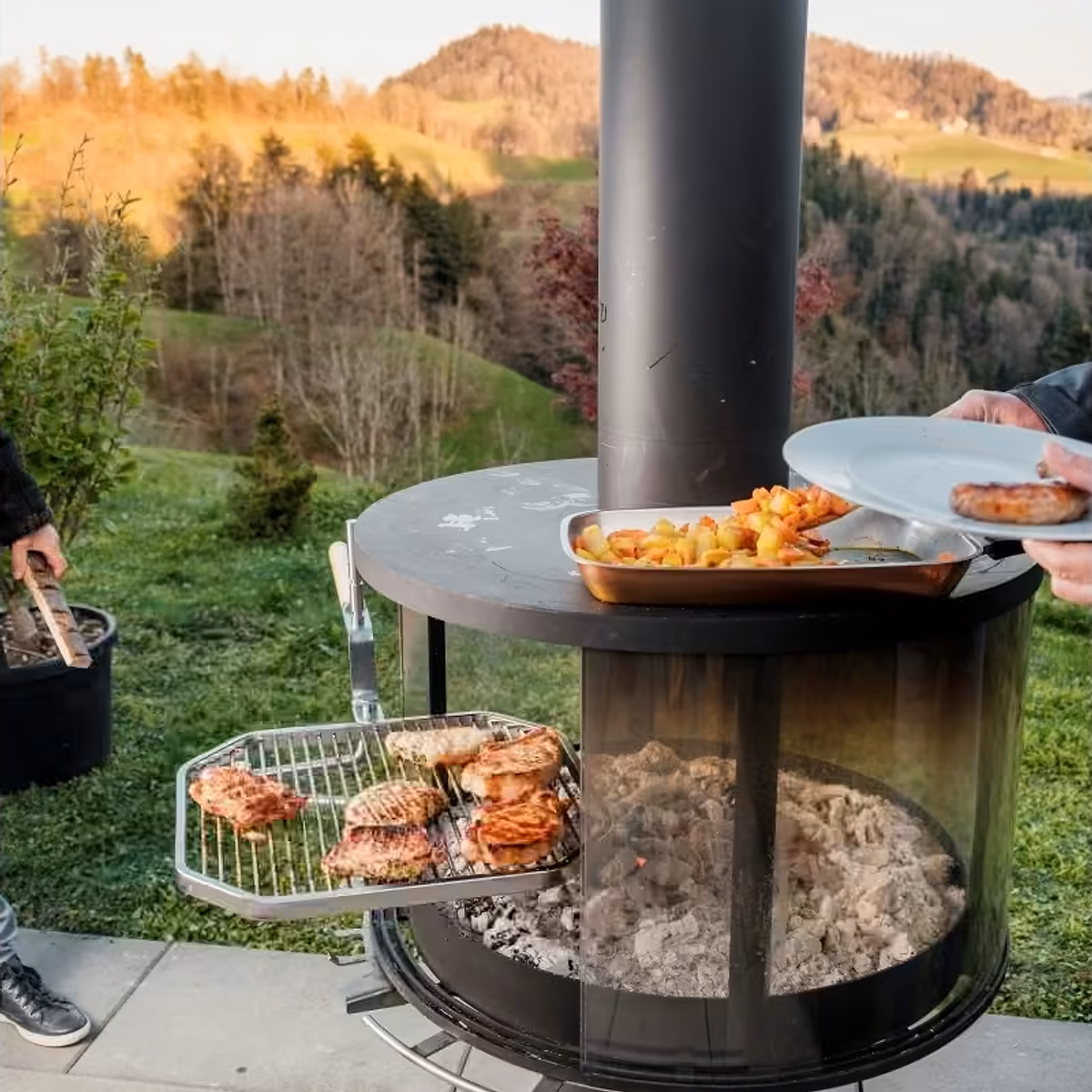 Rüegg Surprise utepeis – folk griller kjøtt og serverer mat på svingbar grill med panoramiske flammer i vakker hage hos Peisbutikken.no