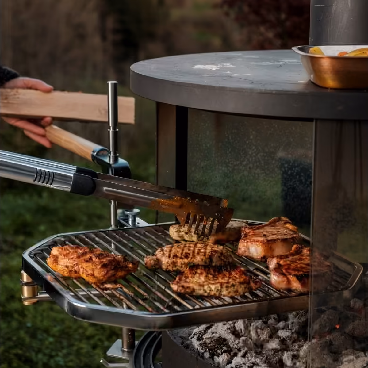 Rüegg Surprise utepeis – nærbilde av grilling kjøtt på svingbar grill med glødende kull og tang i hage hos Peisbutikken.no