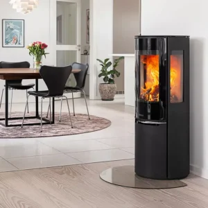 Sidevisning av Aduro 22.5 Lux vedfyrt peisovn i moderne hjem, som viser sort stålramme, panoramaglassdør med oransje flammer, askeboks, nær sofa og hvit vegg, fremhever vedfyrt peis-fleksibilitet.
