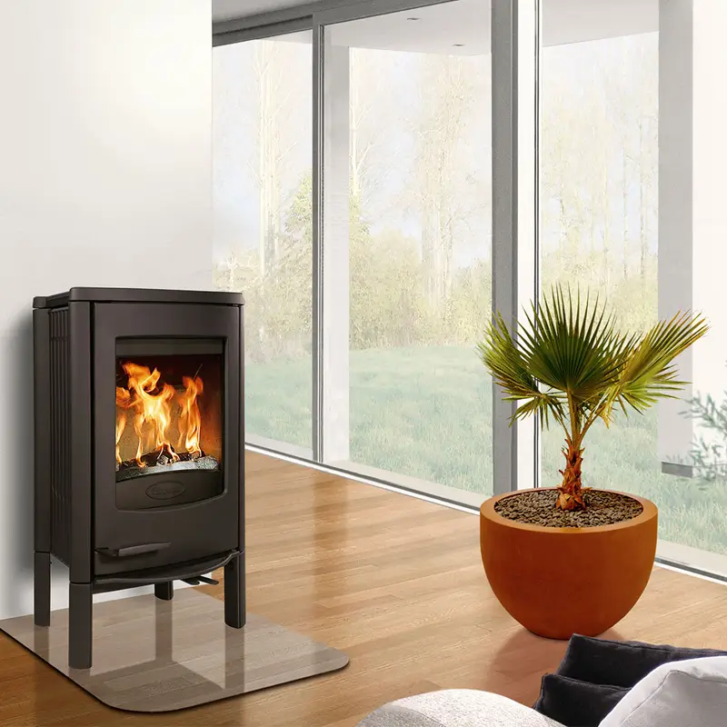 Dovre Cinderella L peisovn lifestyle Dovre Cinderella L vedovn i svart støpejern plassert i et lyst stuehjørne, med stor glassdør som viser flammene, omgitt av tregulv, jordfarger og store vinduer med utsikt til grønn natur.