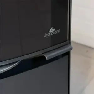 Et nærbilde av Jydepejsen Bella peisovn viser dens blanke, sorte stålskrog med et fremtredende integrert håndtak og et klart glasspanel som viser Jydepejsen-logoen med et flammeikon, satt mot en minimalistisk bakgrunn. Bildet fremhever peisovnens glatte overflate og selvlukkende dør, designet for sikker og effektiv vedfryt-brenning, med en subtil refleksjon som legger dybde til det moderne peis-designet. Basen og håndtaket er skapt med presisjon, og understreker dens holdbare konstruksjon for langvarig peis-varme.