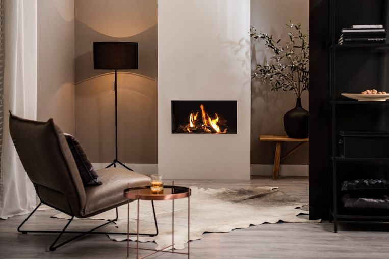 Trimline 73 Front livsstilsbilde – bred moderne innebygd lineær gasspeis med realistiske dansende flammer i elegant nøytral stue med brun skinnlenestol, kobberbord, saueskinnsteppe og høyt svart reolsystem hos peisbutikken.no