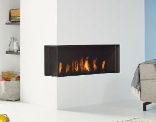 Global 100 Corner BF livsstilsbilde – bred moderne svart hjørne gasspeisinnsats med panoramaglass på to sider og levende realistiske flammer i lys minimalistisk stue med hvite vegger, koselig sofa, hyller og mykt naturlig lys hos peisbutikken.no