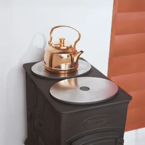 En blank messingkjele står på kokeplaten til en svart vedovn i støpejern. De runde metallplatene på toppen er laget for å varme vann eller tilberede enkle måltider.