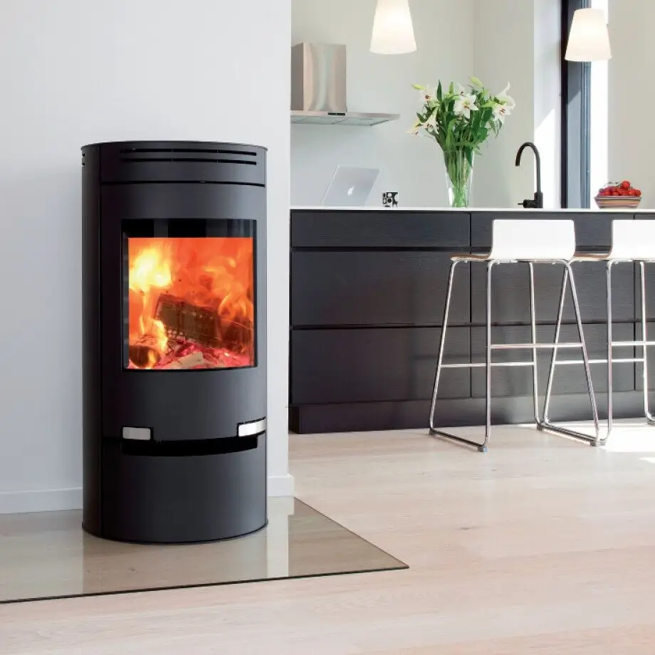 Sort sylindrisk Aduro 1.1 peisovn med levende oransje flammer synlige gjennom dens store buede glassdør, som viser en solid støpejernskonstruksjon og glatt platejernsfinish, plassert på et lyst tregulv i et moderne kjøkkenmiljø med elegante sorte skap, hvite barkrakker, en vase med hvite liljer på benken, en sølvkran, friske frukter i en bolle, en laptop og pendellamper overhead, som understreker peisovnens kompakte fotavtrykk og elegante designintegrasjon for peis og vedfyrt bruk.