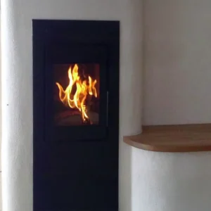 Bilde av Rüegg KE 36x74x48 peisinnsats montert på vegg, med matt sort ramme og stort glassfelt som viser levende flammer. Omgitt av lyse vegger, en buet hvit flate og en flytende trehylle som gir et moderne og harmonisk uttrykk.