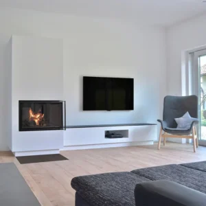 Rüegg KII 50x68x50 tosidig peisinnsats med front- og høyreglass, flush-montert i en hvit vegg i en moderne stue med lyst tregulv og skandinavisk interiør.