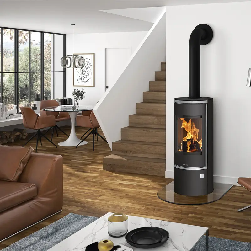 Lifestyle-bilde av Oranier Corso 2.0 Sort Stål peisovn i et moderne, åpent oppholdsrom, plassert ved siden av en trapp. Den høye, runde kroppen i sort stål og den store glassdøren viser flammene, omgitt av varmt tregulv, moderne møbler og store vinduer som slipper inn naturlig lys.