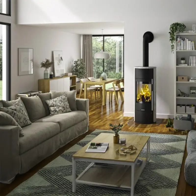 Oranier Polar Neo Vantage W+ peisovn med klebersteinsfinish står sentralt plassert i et lyst og innbydende stuerom. Den slanke, svarte, sylindriske formen og den levende flammen skaper en varm kontrast mot den lyse bakveggen. Rundt ovnen finnes en beige sofa, et trebord med bøker, og stilfull dekor i et koselig skandinavisk interiør badet i naturlig lys.