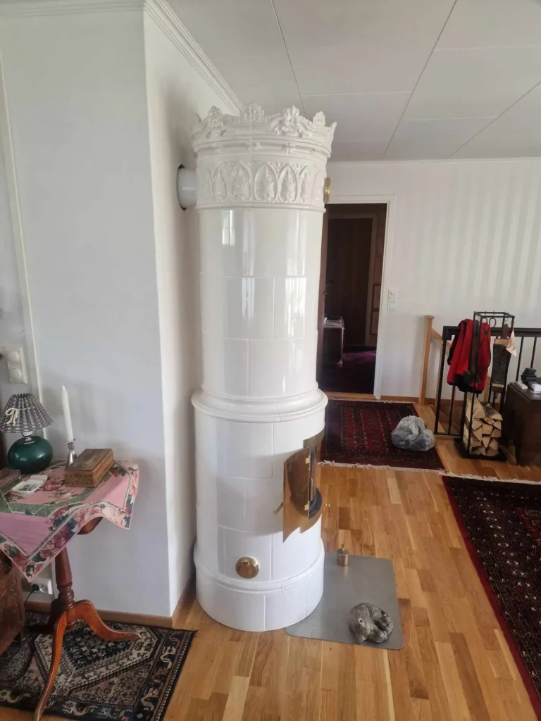 Klassisk hvit kakkelovn med ornamentert topp i koselig stue med tregulv og vedkurv – profesjonell montering av tradisjonelle peiser fra Peisbutikken.no