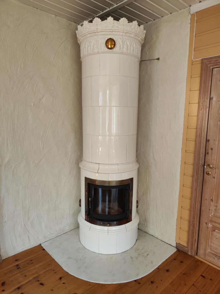 Elegant kakkelovn montert av Peisbutikken i Enebakk – klassisk hvit kakelovn med ornamentert krone og buet glassdør i koselig hjørne